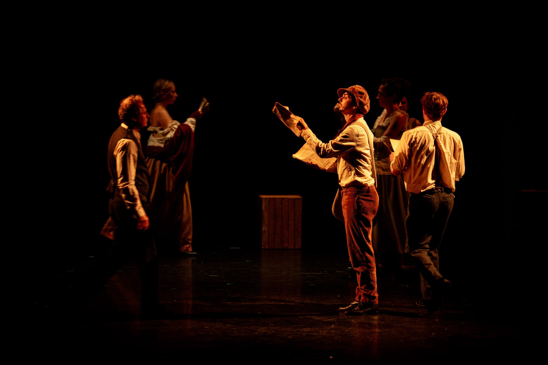 Un comédien lit un journal sous une lumière ambrée, entouré de la troupe
